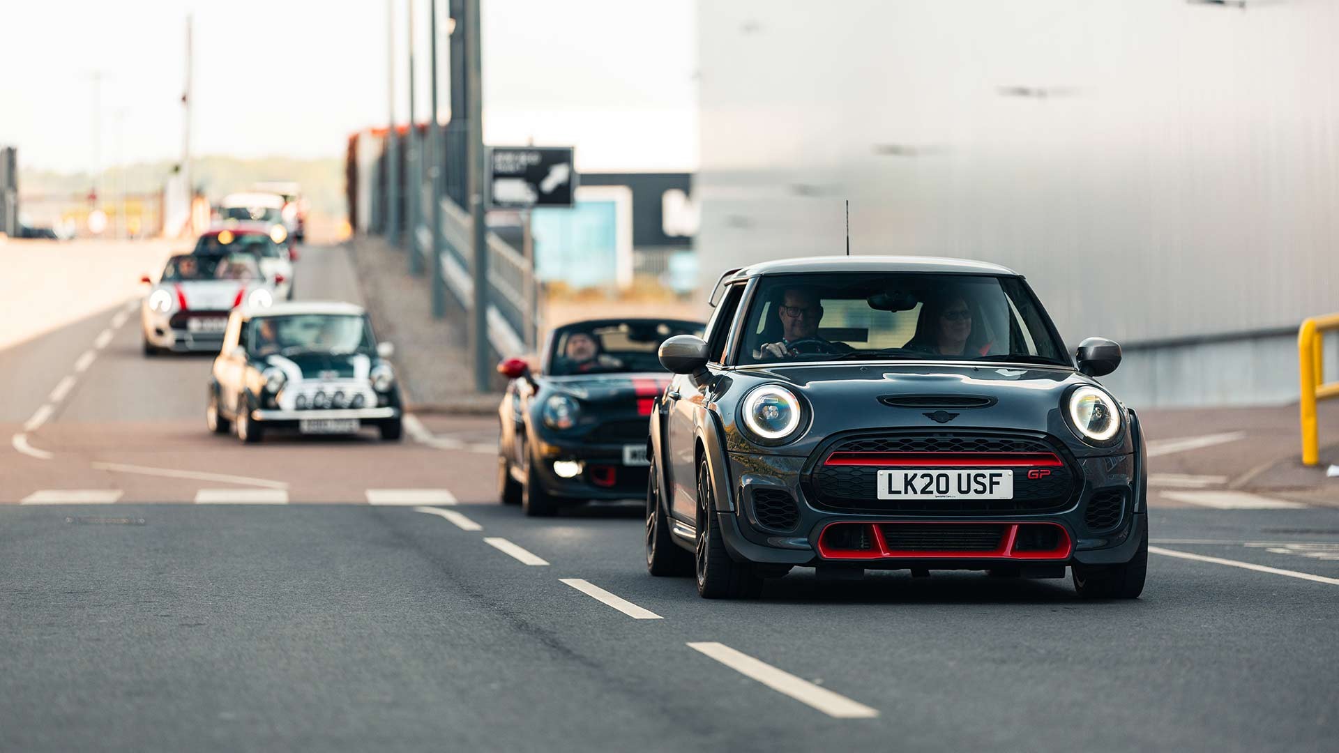 A line of Mini Coopers, led by a black one with red accents, drives down a road near industrial buildings.  